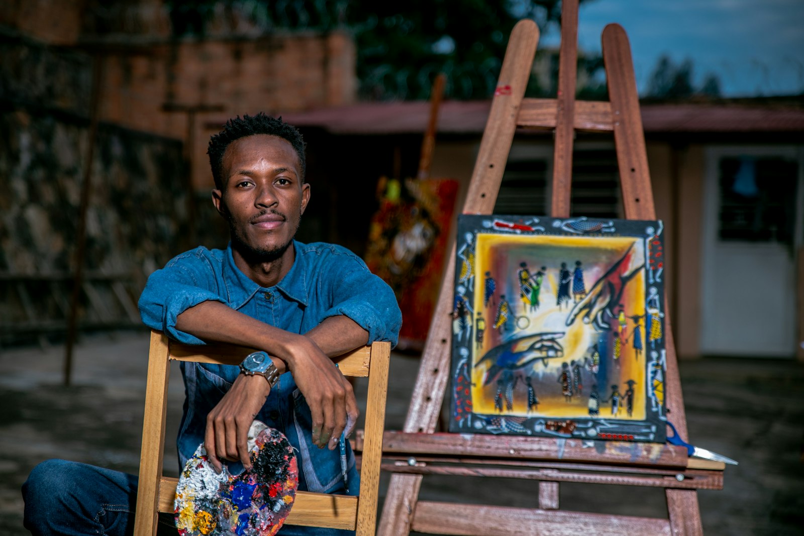 A painter with a loaded palette beside a finished canvas on an easel